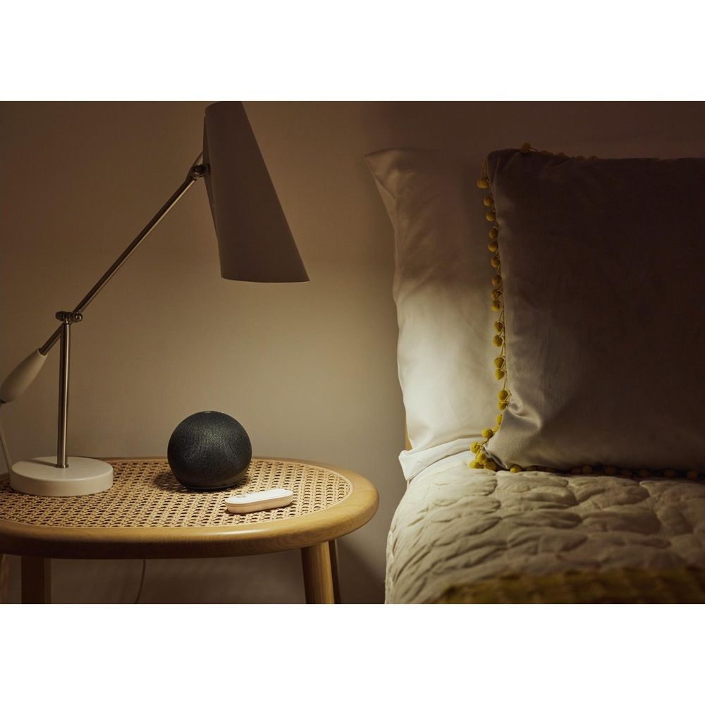 Una mesita de noche con lámpara, altavoz negro y mando junto a una cama.
