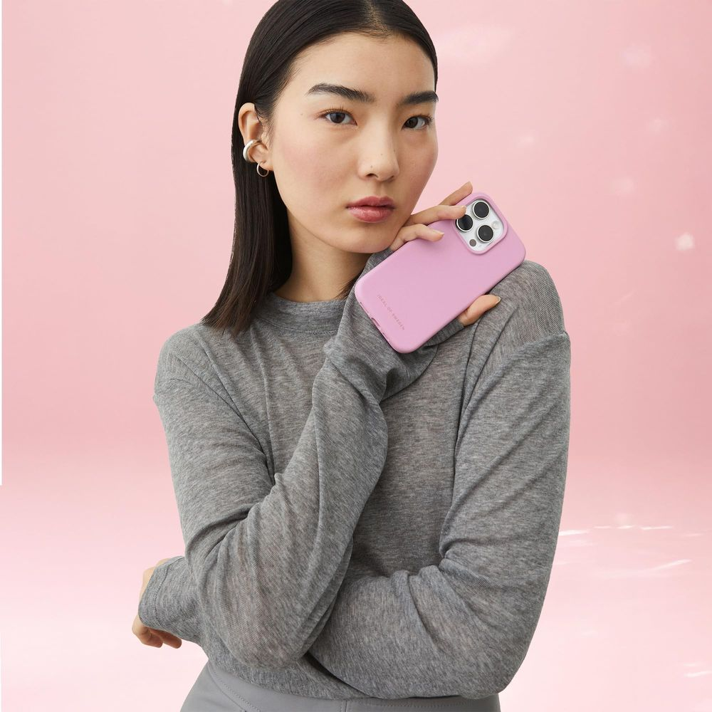 Eine Frau mit grauer Kleidung hält ein rosafarbenes Handy vor einem rosa Himmel.
