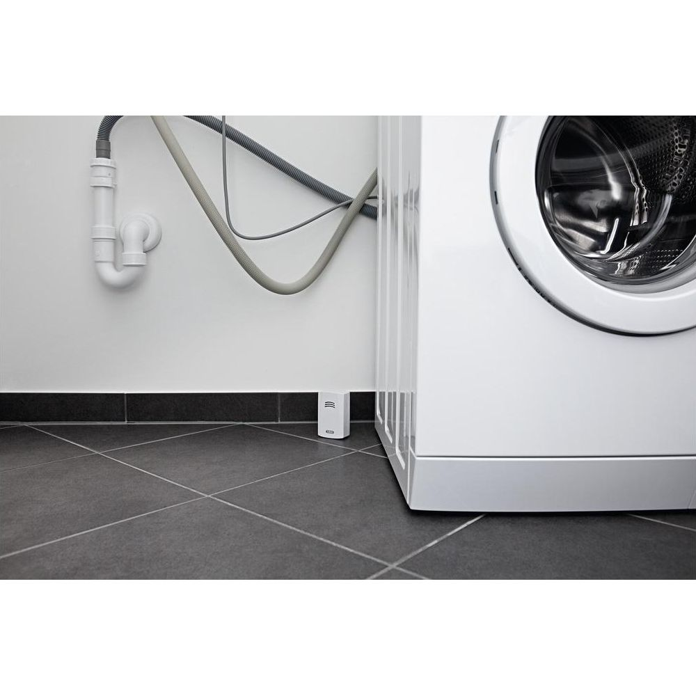 Ein kleiner weißer Sensor mit Wellenlinien steht auf dunkelgrauem Boden neben einer Waschmaschine.