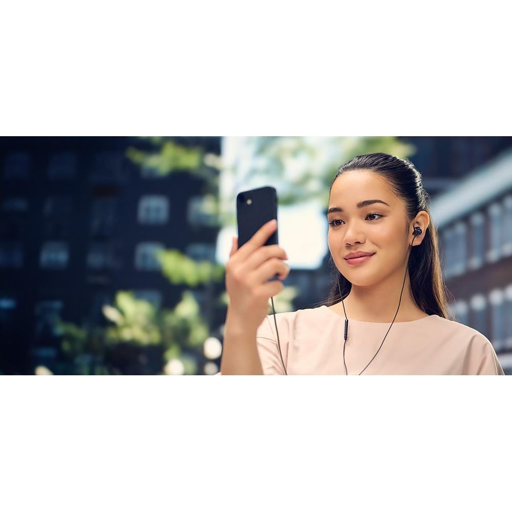 Junge Frau macht Selfie mit Smartphone und Ohrhörern, im Hintergrund Gebäude.