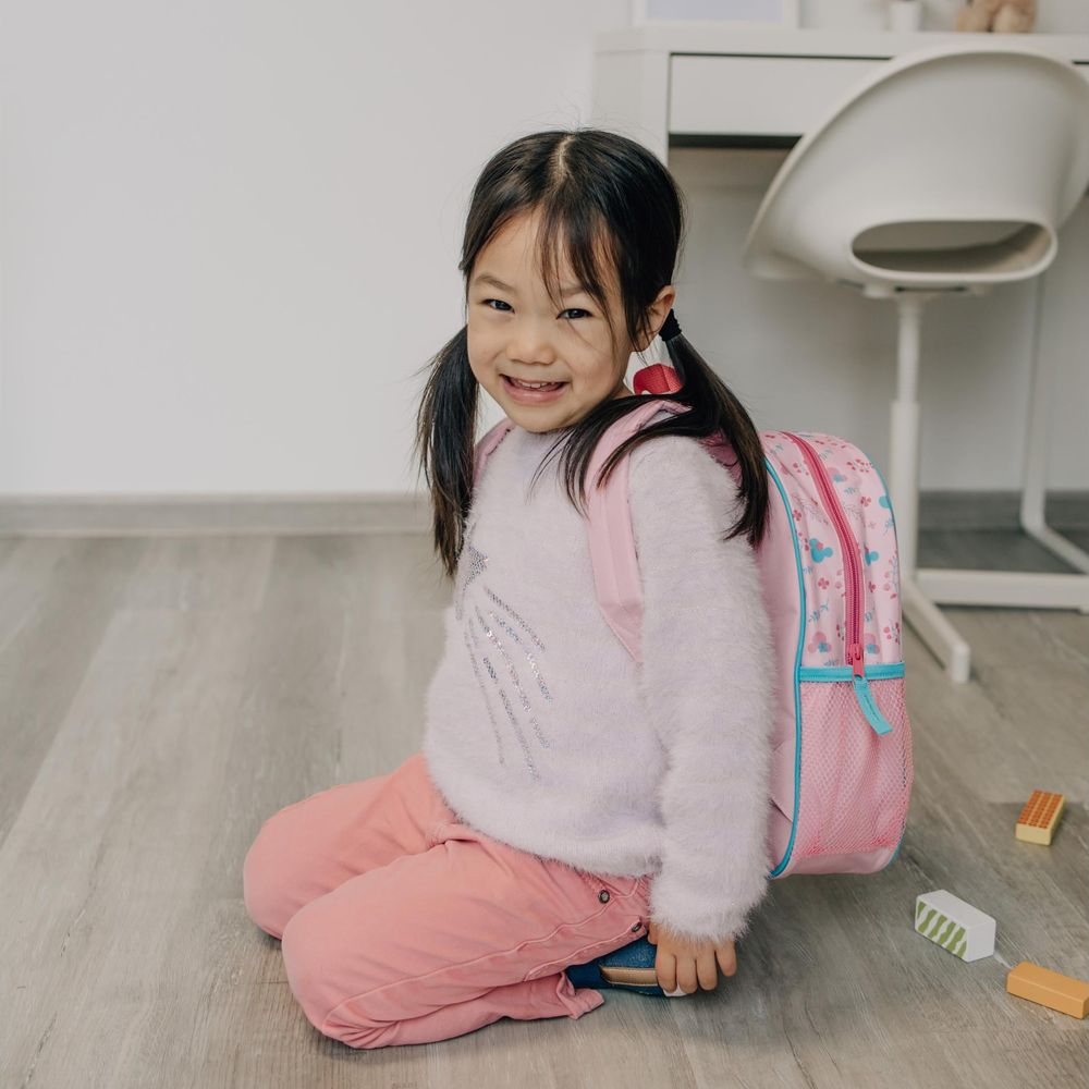 A young Asian girl with pigtails sits on the floor wearing a pink backpack.