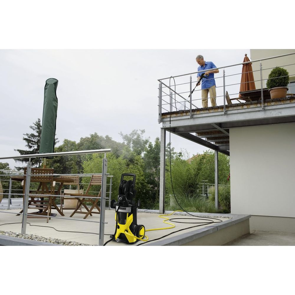 Mężczyzna czyści balkon myjką ciśnieniową; żółta maszyna poniżej.