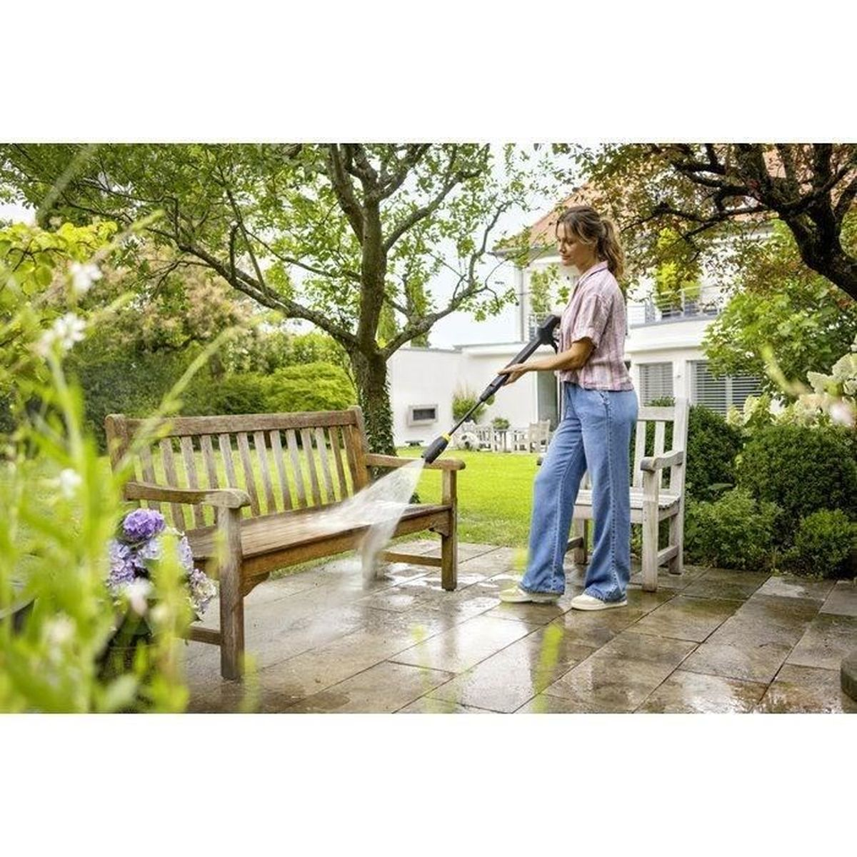 Kobieta czyści drewnianą ławkę myjką ciśnieniową w ogrodzie.