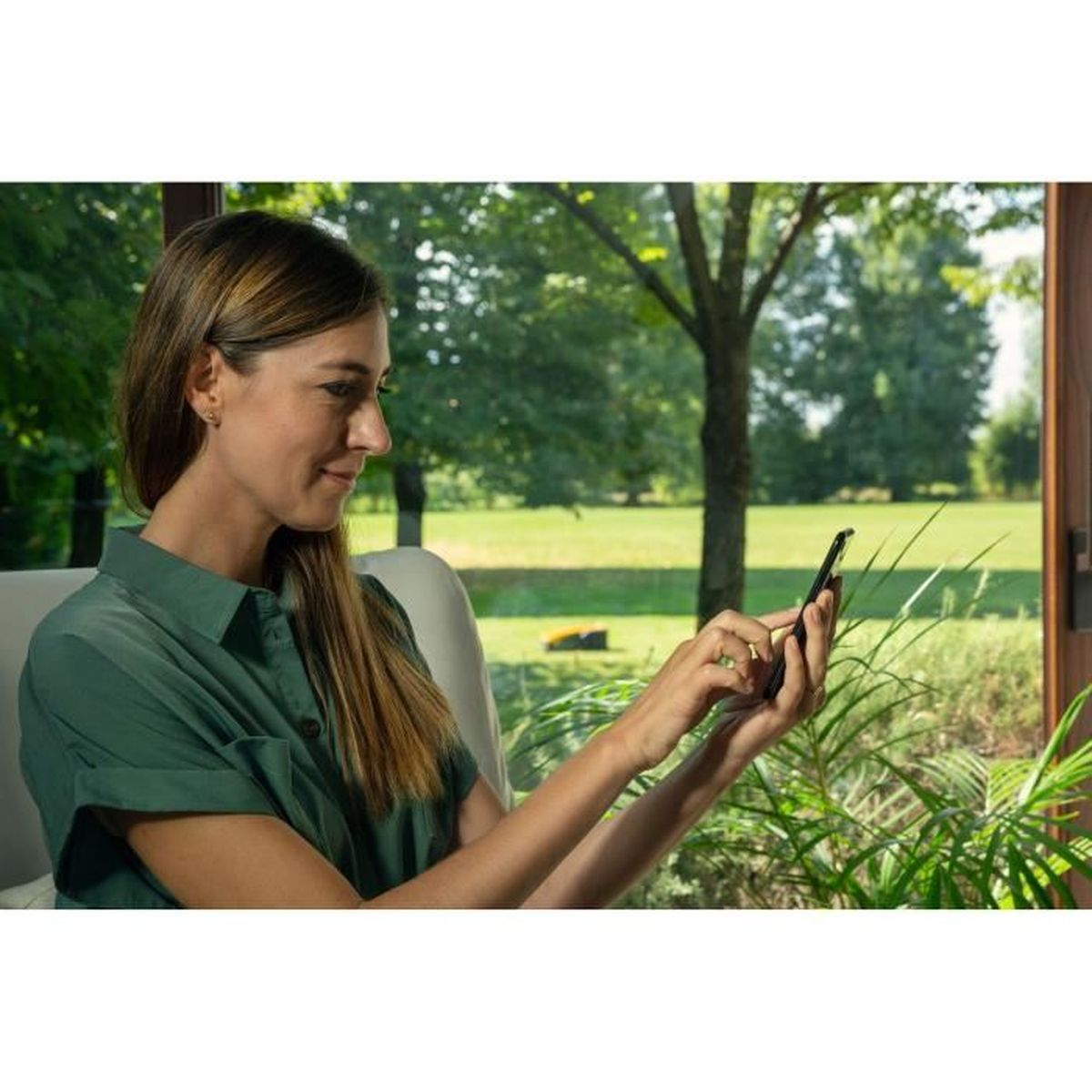 Mujer con camisa verde usa teléfono adentro. Cortacésped robot afuera.
