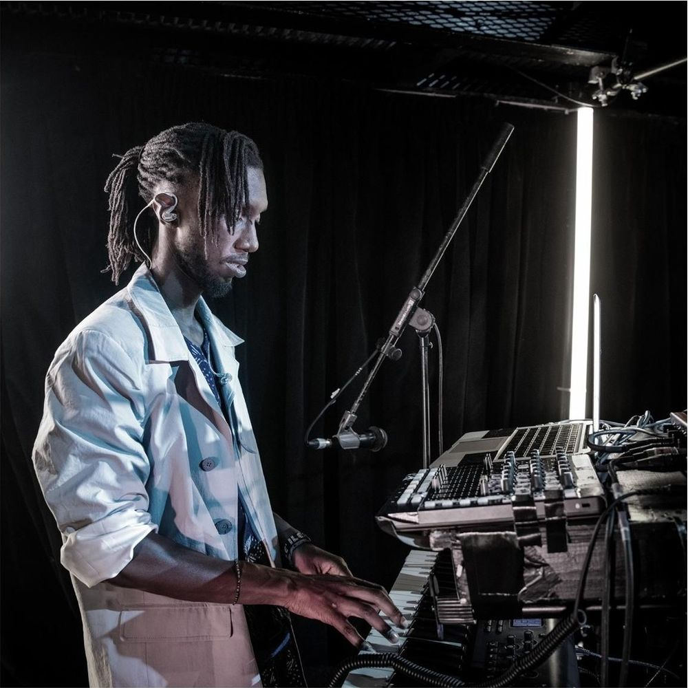 Mężczyzna z dredami gra na keyboardzie przed czarną kurtyną, oświetlony neonami.