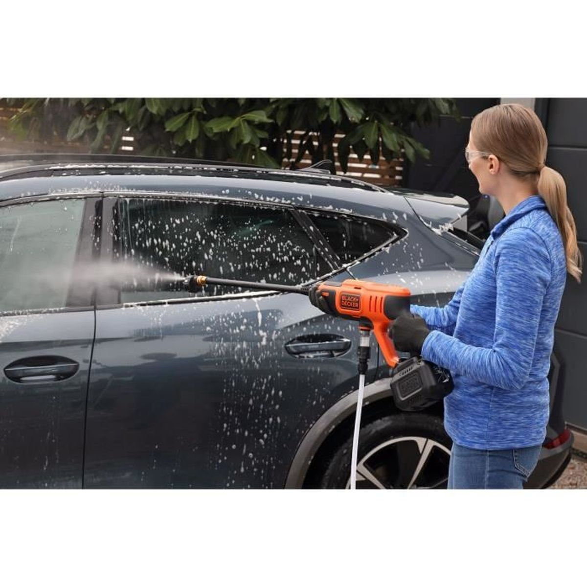 Eine Frau mit Brille wäscht ein dunkles Auto mit einem Hochdruckreiniger.