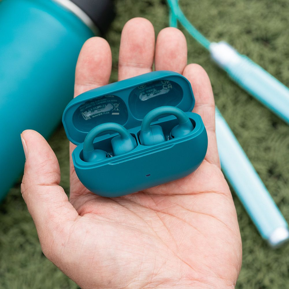 Turkusowe słuchawki w etui ładującym trzymane w dłoni, z butelką wody.