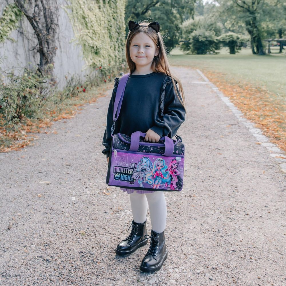Ein junges Mädchen mit langen braunen Haaren und Katzenohren-Stirnband, einem schwarzen Oberteil und einer weißen Strumpfhose, hält eine lila Monster High Tasche und steht auf einem Weg.