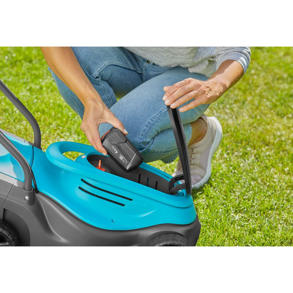 Person setzt Akku in einen blauen Rasenmäher auf grünem Gras.