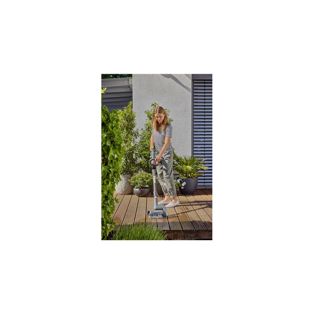 Femme nettoie terrasse en bois avec un appareil, entourée de plantes.