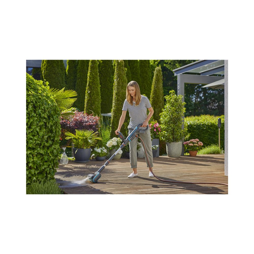 Une femme nettoie une terrasse en bois avec un nettoyeur à brosse et un tuyau.