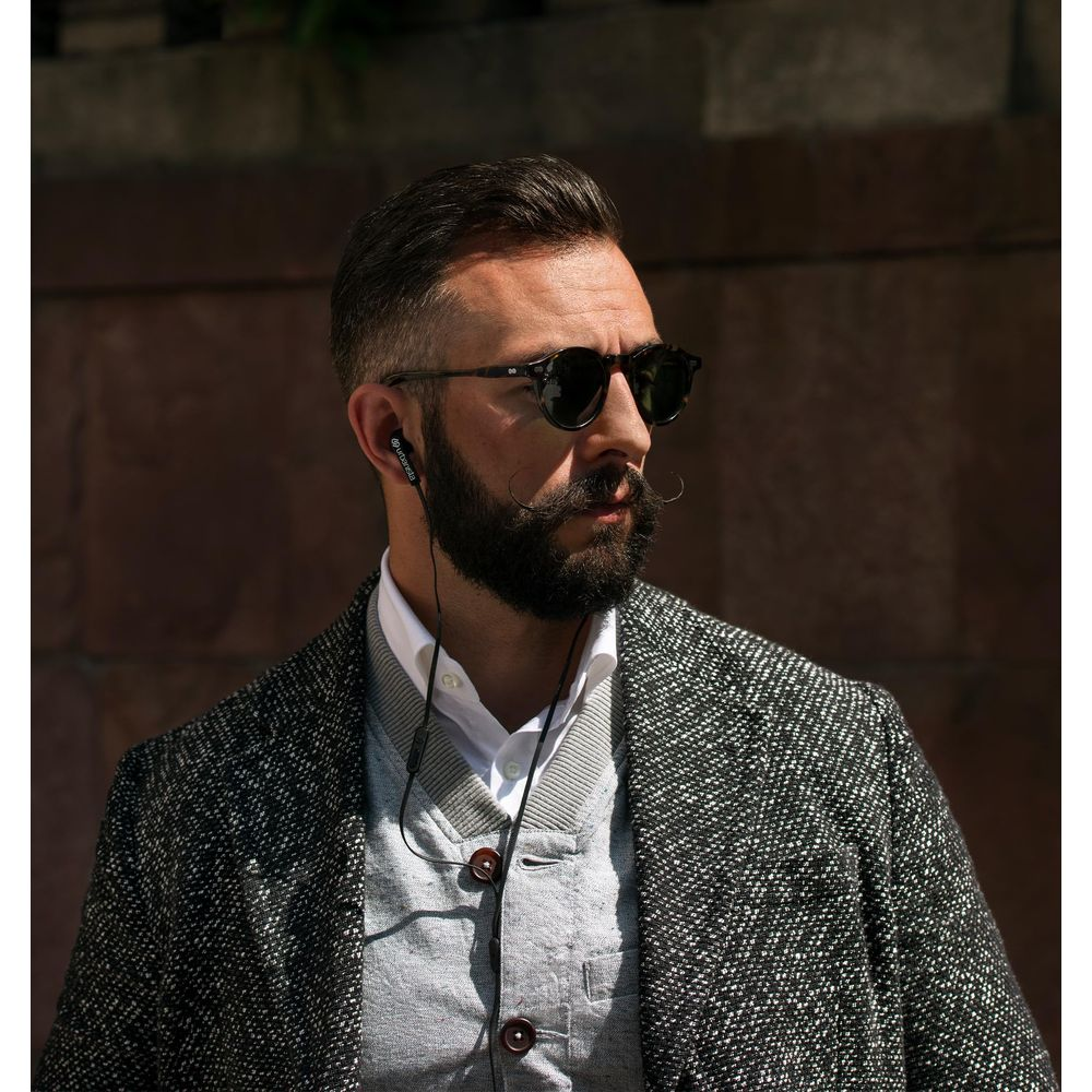 Hombre con barba y bigote usa gafas de sol, audífonos y chaqueta.