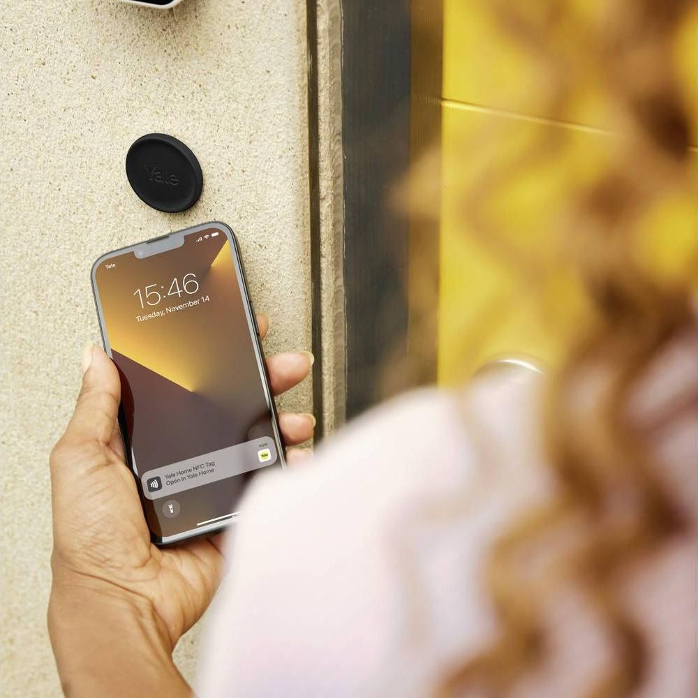 Una mano sostiene un smartphone cerca de una pared con una etiqueta Yale. La hora es 15:46.