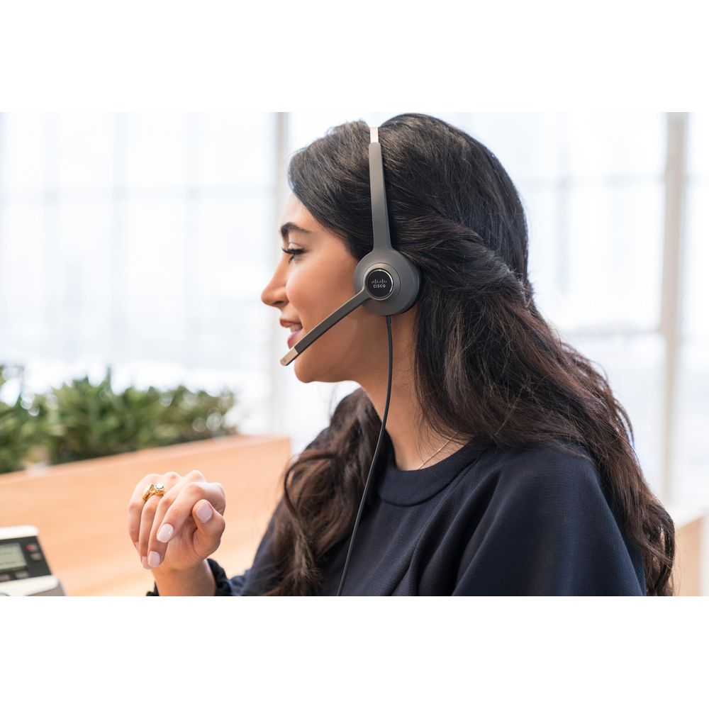 Una mujer con auriculares habla, con el dedo levantado. Plantas y un escritorio al fondo.