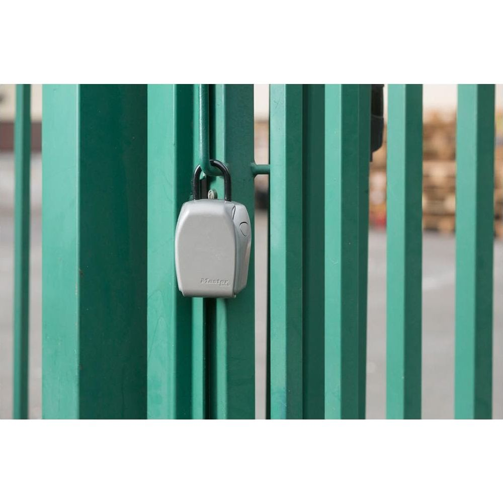 Un candado plateado para llaves cuelga de una valla de metal verde.