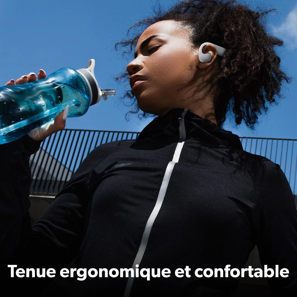 Une femme boit une bouteille d'eau bleue, porte des écouteurs et un haut noir.