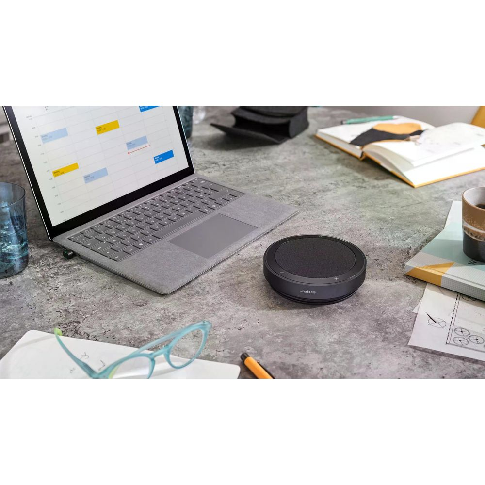 Laptop, głośnik Jabra i okulary na biurku.
