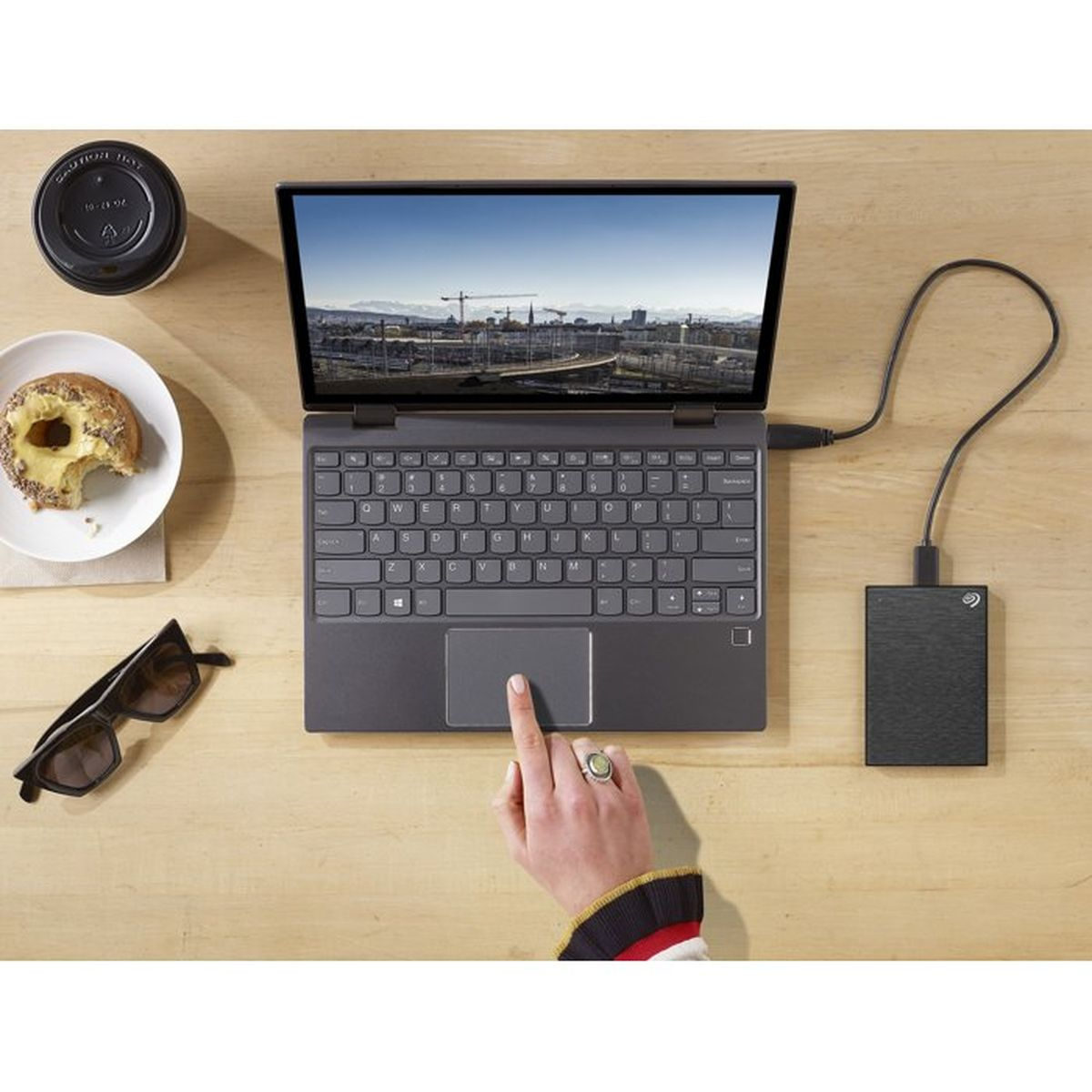 Laptop z zewnętrznym dyskiem twardym, kubek kawy, pączek i okulary przeciwsłoneczne.