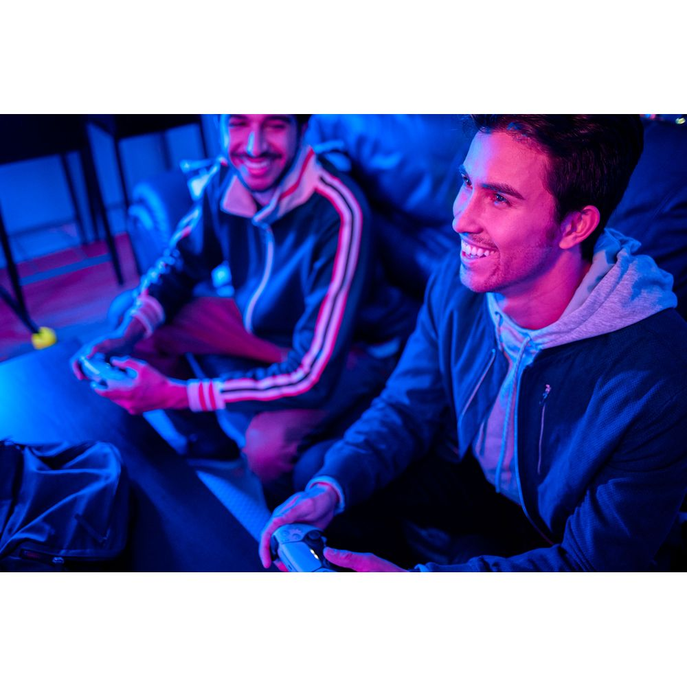 Dwóch uśmiechniętych młodych mężczyzn gra w gry wideo w ciemnym pomieszczeniu oświetlonym niebieskim światłem.