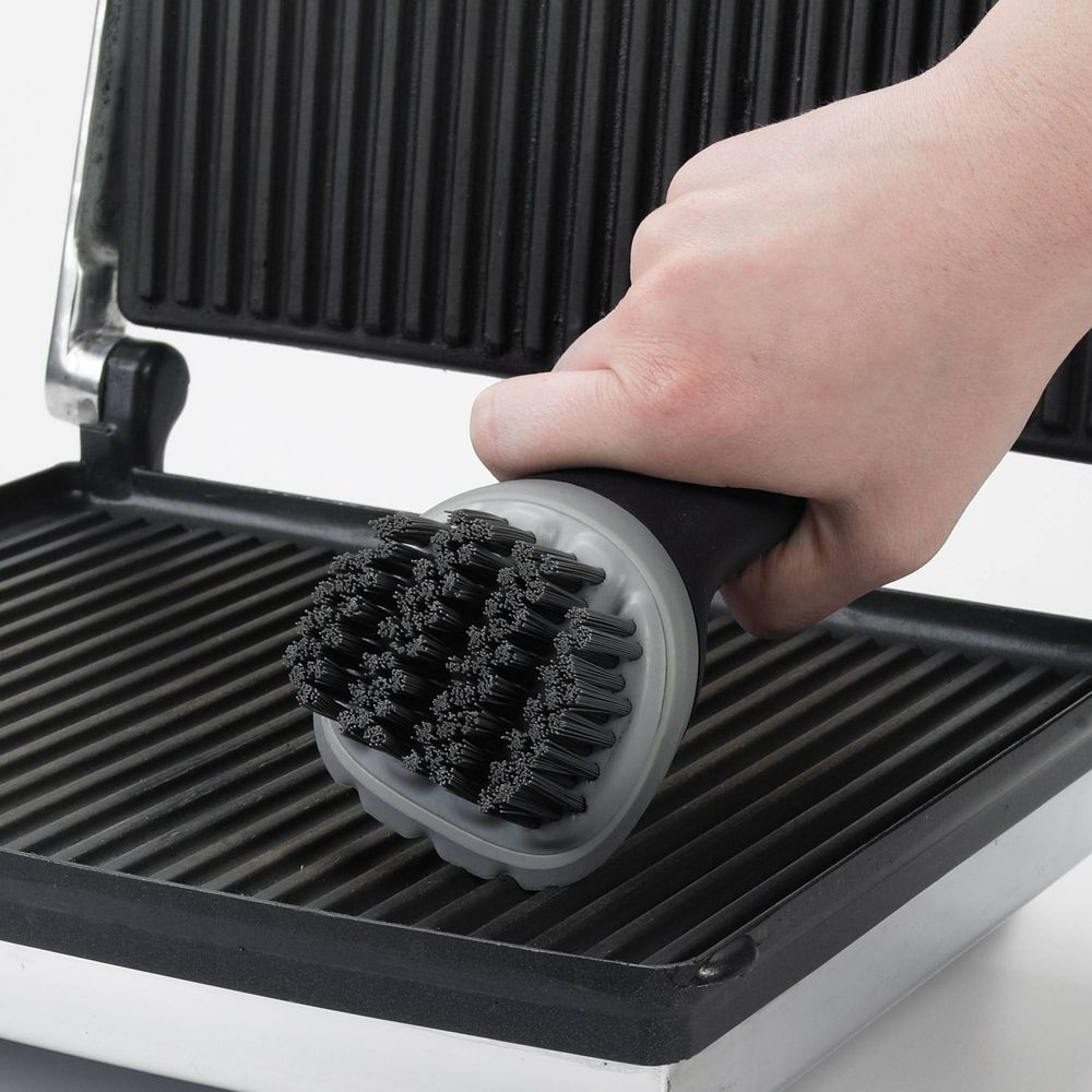Hand reinigt Grill mit Bürste. Schwarze Borsten auf gerillter Oberfläche.