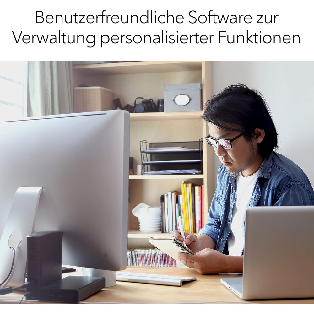 Mężczyzna siedzi przy biurku, pisząc w notatniku. Widoczny komputer i laptop.