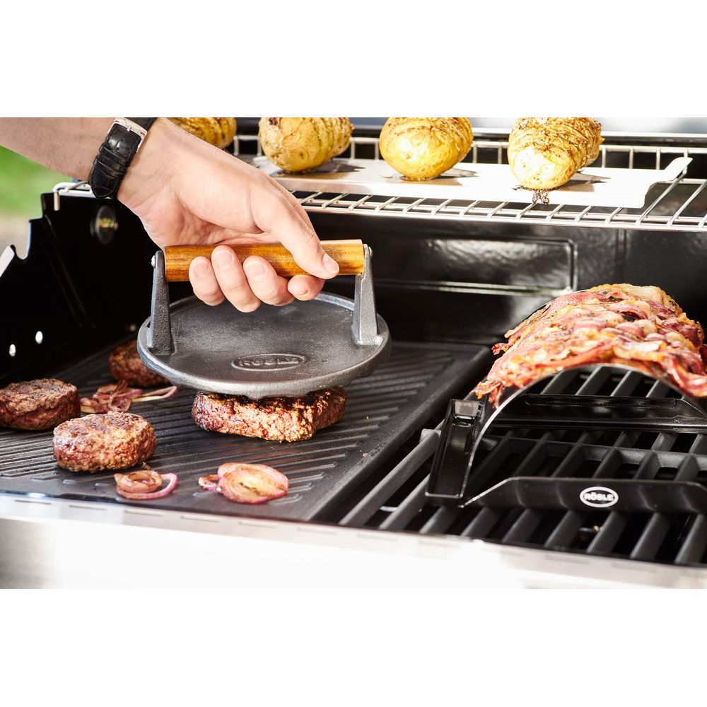 Hand drückt Burgerpresse auf Pattys auf dem Grill. Zwiebeln und Kartoffeln daneben.