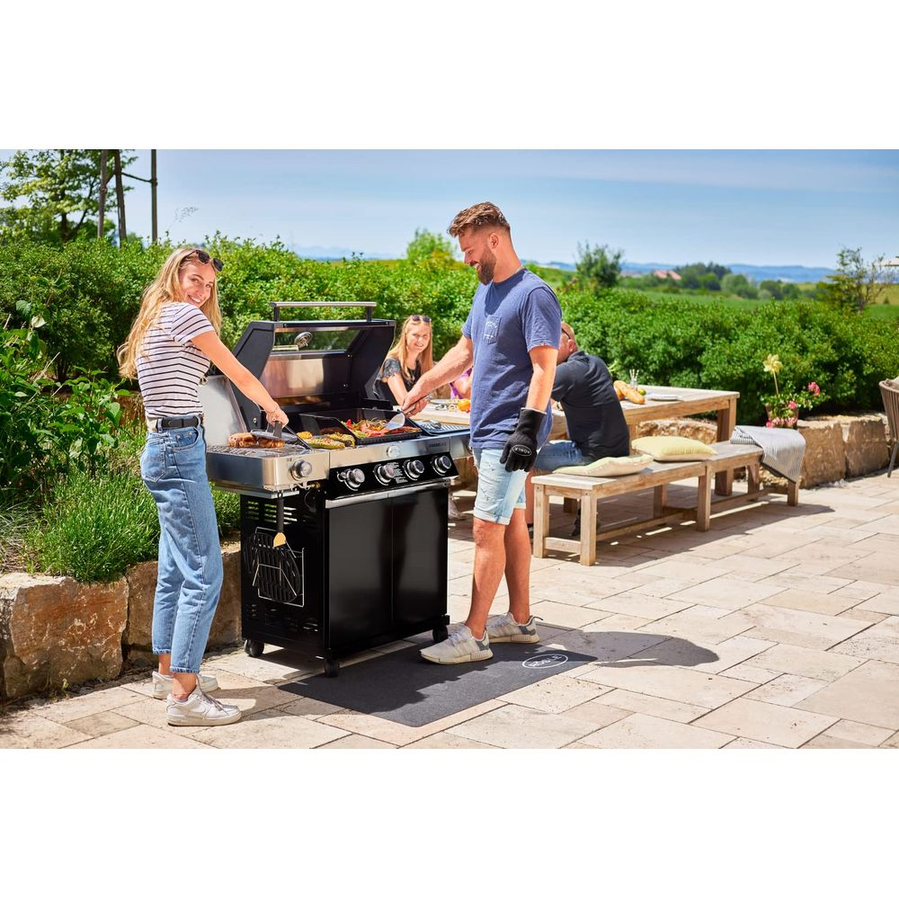 Ein Mann und eine Frau grillen im Freien. Andere sitzen an einem Tisch.
