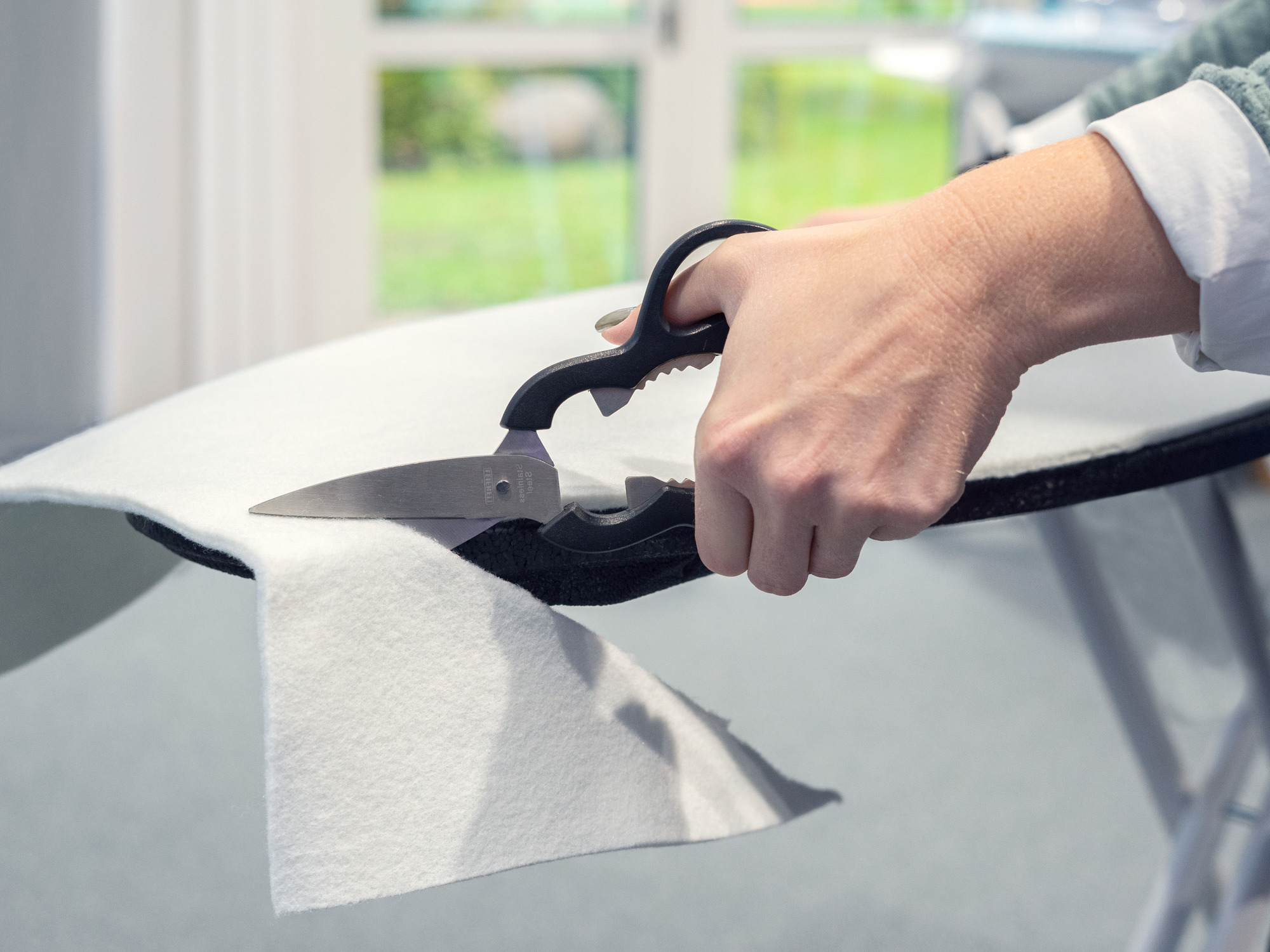 Dłoń tnie biały filc nożyczkami na desce do prasowania.