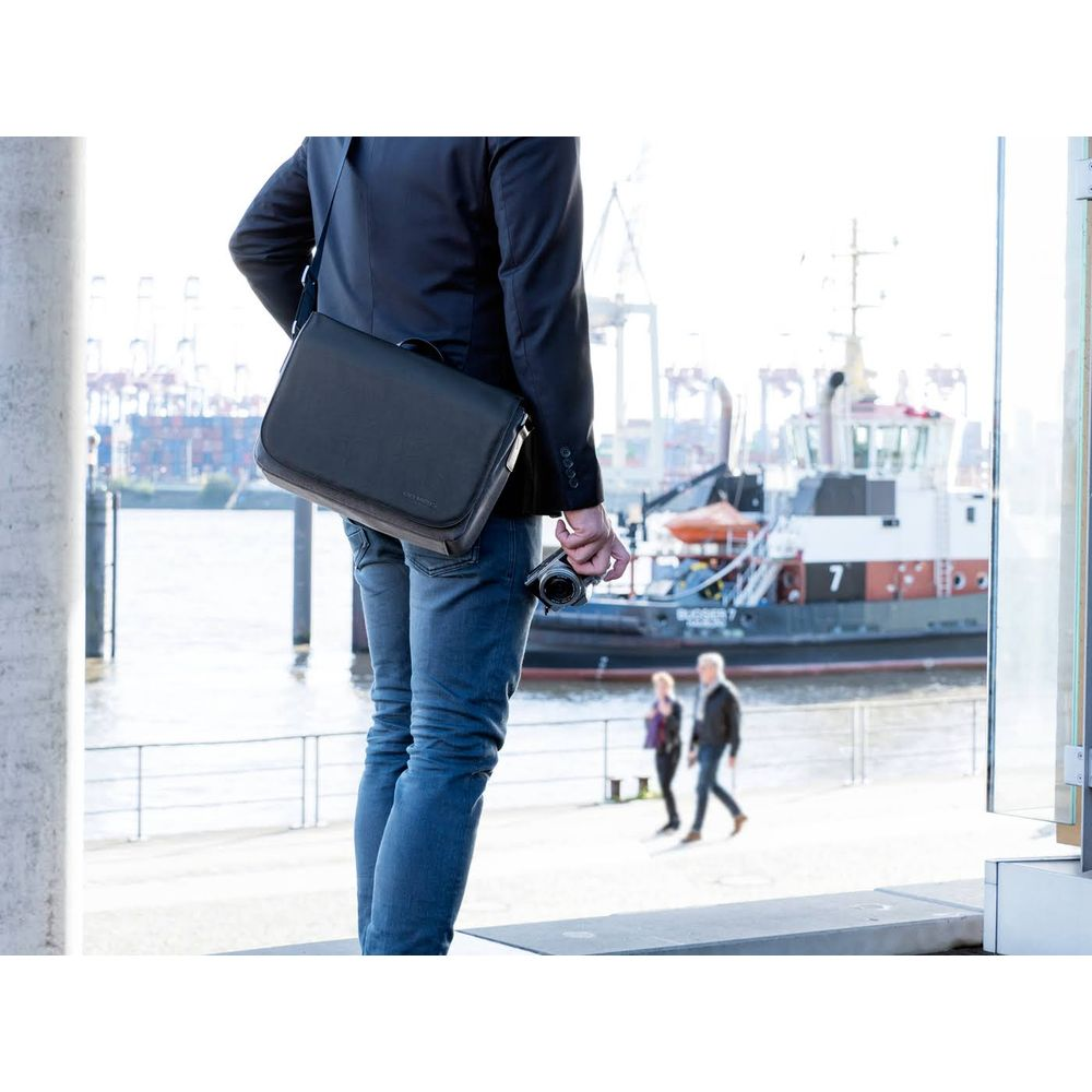 Uomo con borsa nera e macchina fotografica, porto sullo sfondo con barche e persone.