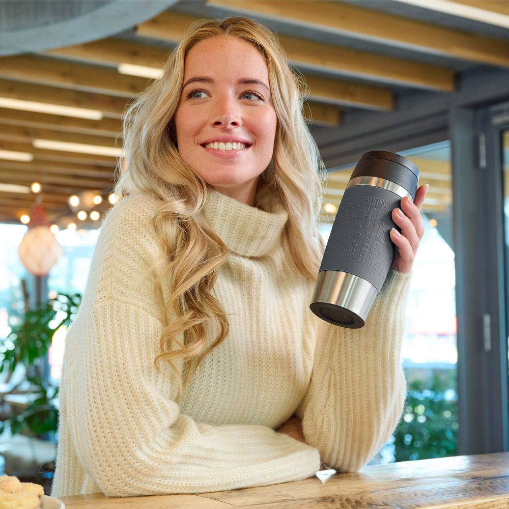 Eine Frau in einem beigen Pullover hält einen Thermobecher in der Hand.