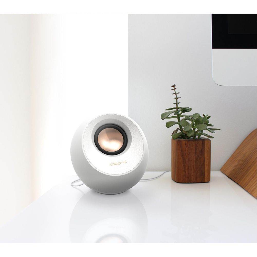 Biały, kulisty głośnik z miedzianym środkiem stoi na białym biurku obok rośliny w doniczce.