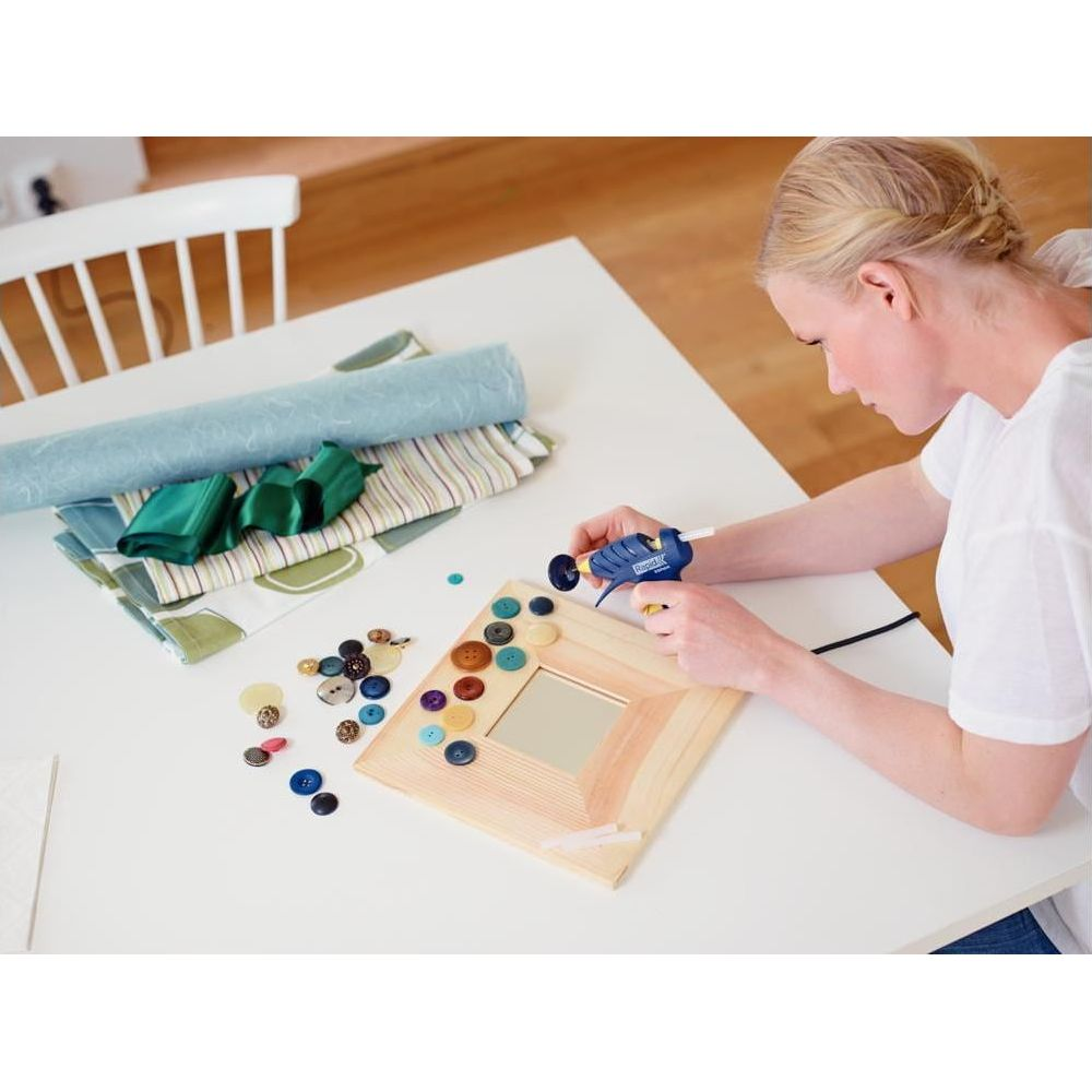 Kobieta przykleja guziki do drewnianej ramki pistoletem na klej.