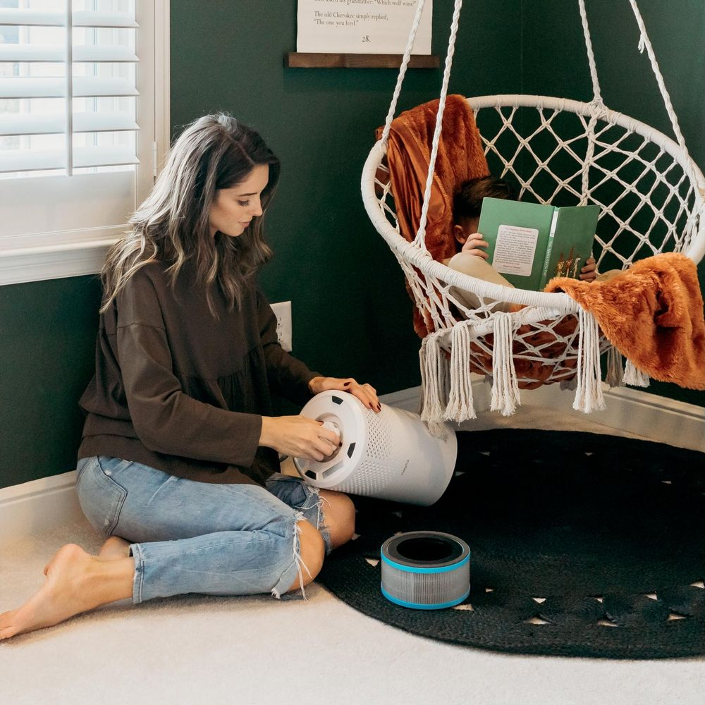 Kobieta siedzi na podłodze, wymieniając filtr oczyszczacza powietrza. Dziecko czyta w wiszącym fotelu.