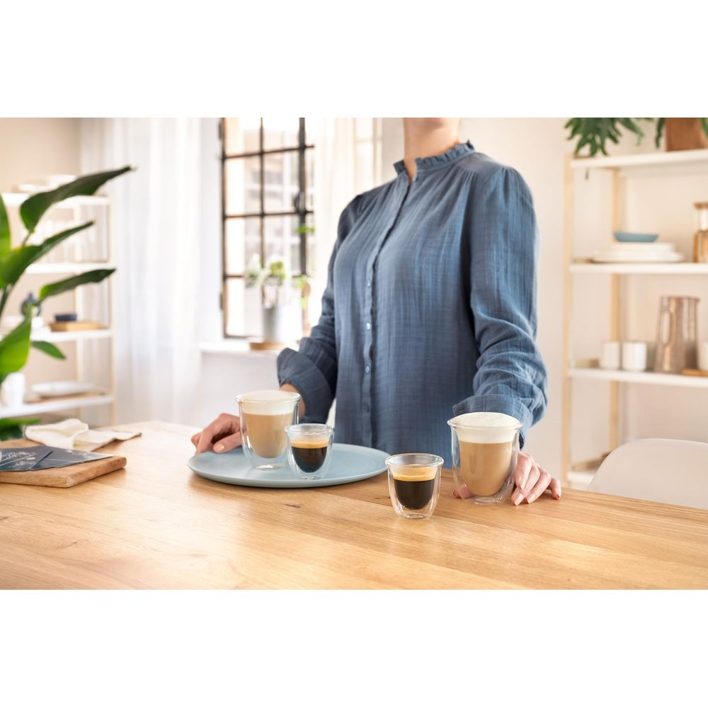 Una persona con camisa azul presenta tres vasos de café en una mesa de madera.