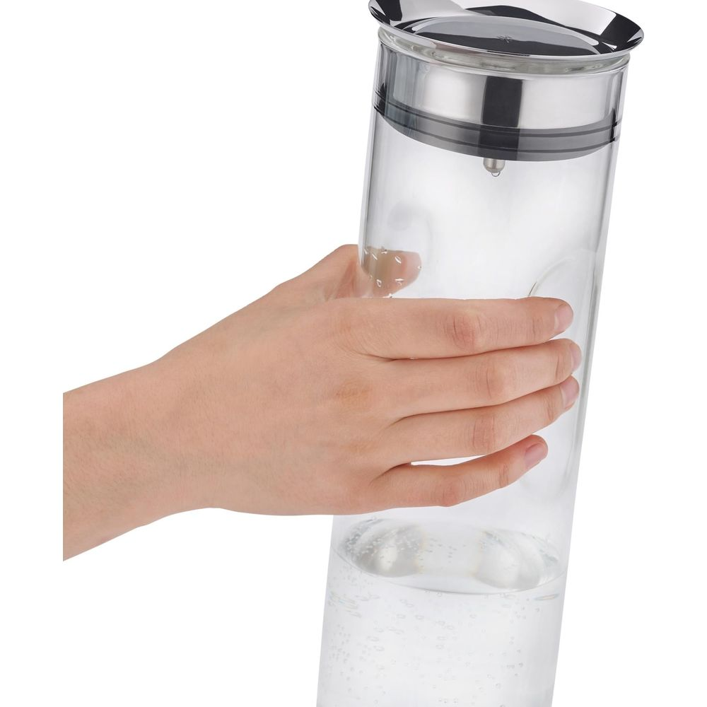 Eine Hand hält einen Glas-Wasserkrug mit einem silbernen Deckel. Wasser ist im Krug.