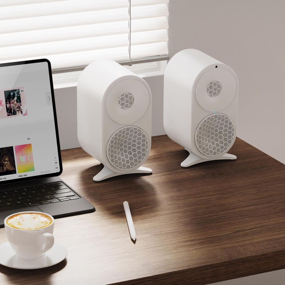 Dos altavoces blancos en un escritorio con portátil, taza y lápiz.