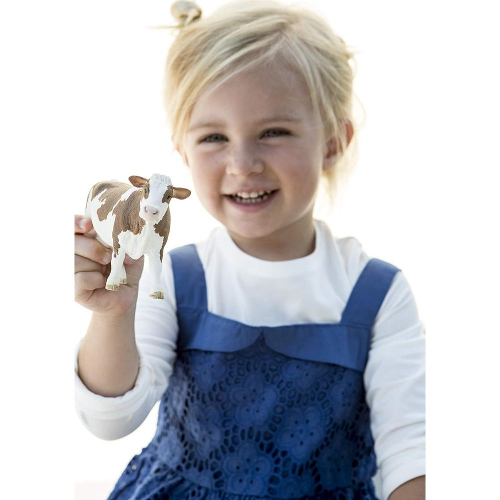 Uśmiechnięta dziewczynka z blond włosami w niebieskiej sukience trzyma biało-brązową zabawkę krowę na jasnym tle.