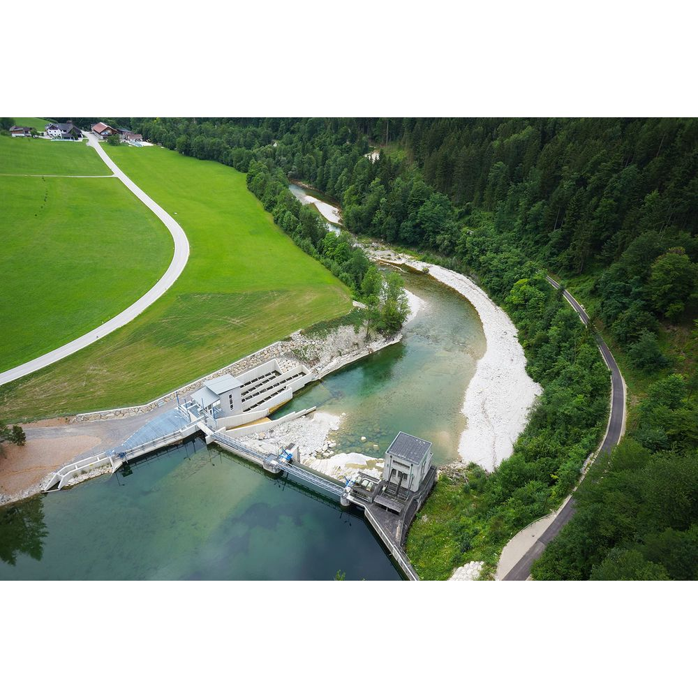Luftaufnahme eines Wasserkraftwerks an einem Fluss, mit grünen Feldern und Wald.