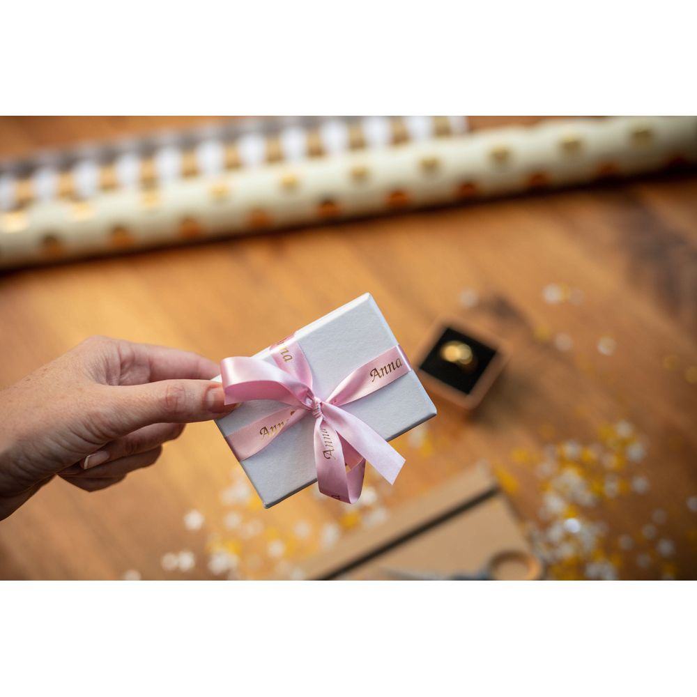 Eine Hand hält eine Geschenkbox mit rosa Band. Ein Ring liegt daneben.
