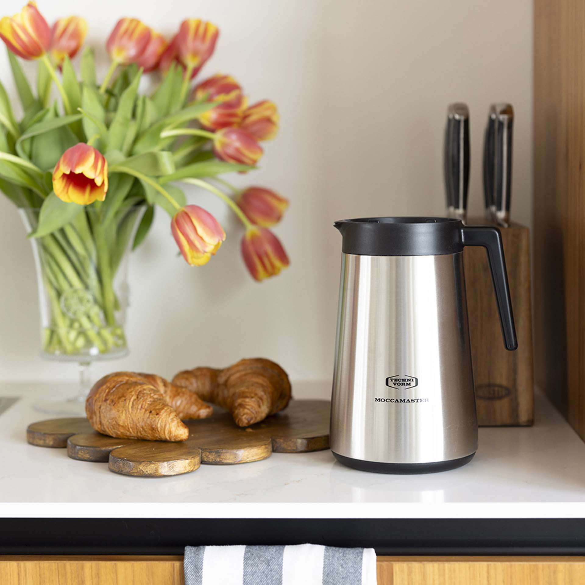 Ein silberner Thermosbehälter steht neben Tulpen und Croissants.