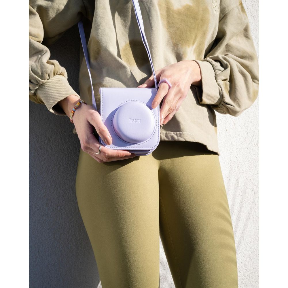 Una persona sostiene un estuche de cámara Instax lavanda con correa morada, vistiendo sudadera tie-dye y pantalones verdes.
