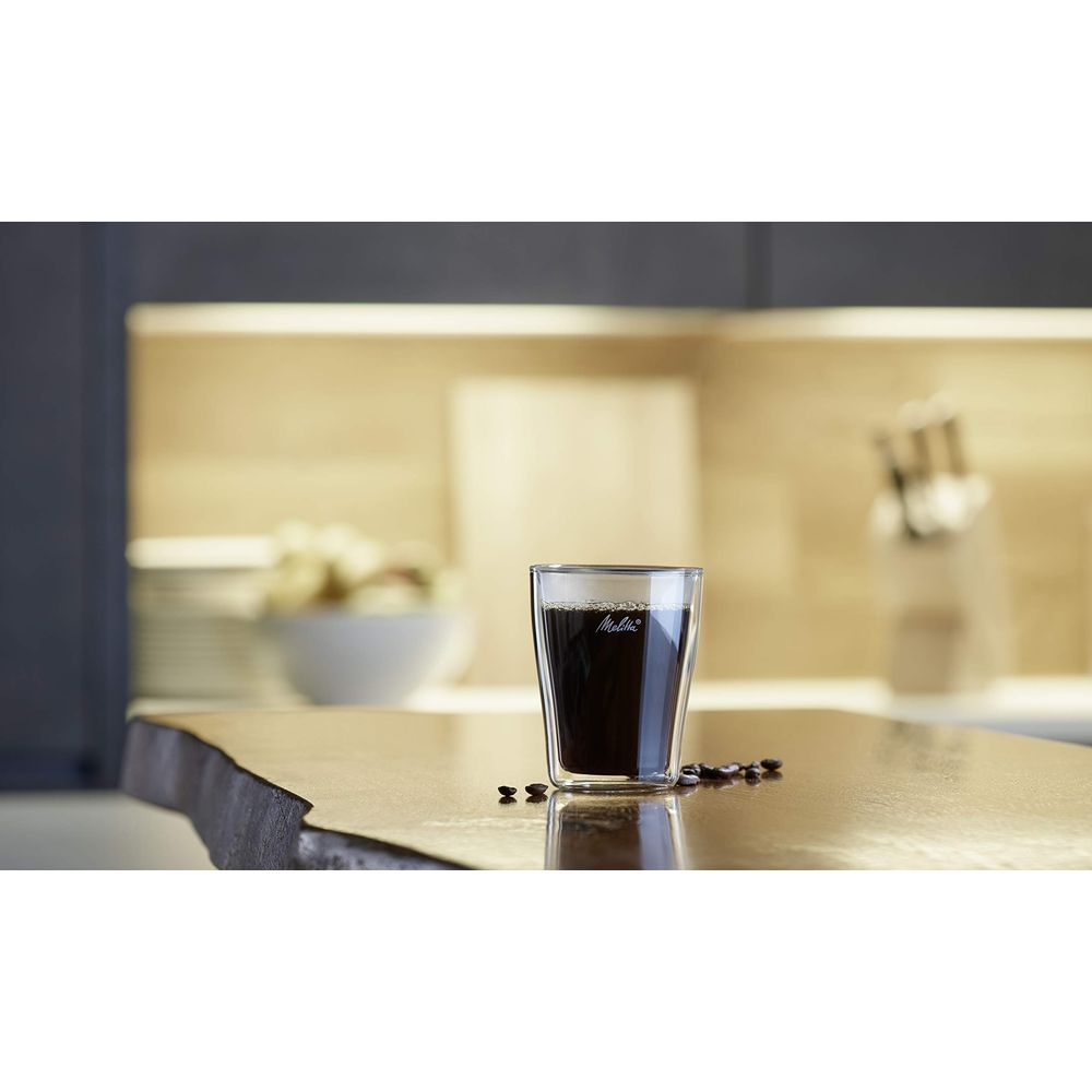 Ein Glas Kaffee mit Bohnen auf einem Holztisch, Küche im Hintergrund.