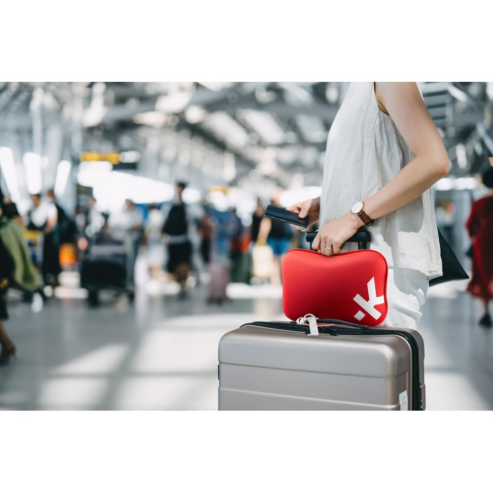Une personne tient un sac rouge avec un K blanc et une valise argentée dans un aéroport animé.
