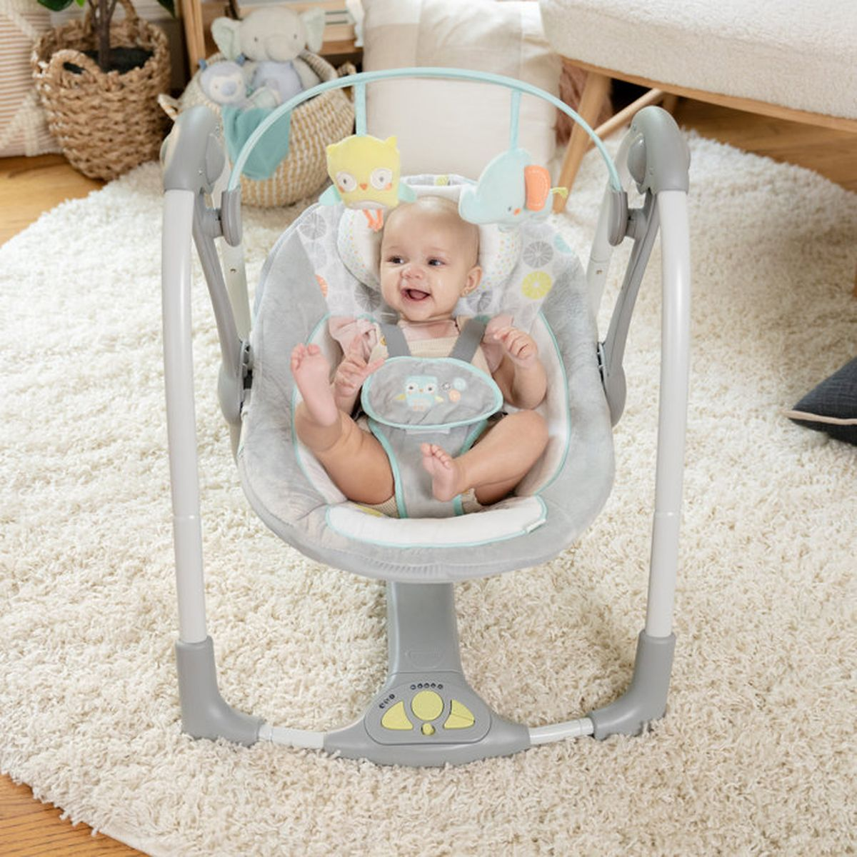 Ein Baby in einem grauen Schaukelstuhl mit Eulenmotiv, sitzend auf einem cremefarbenen Teppich.