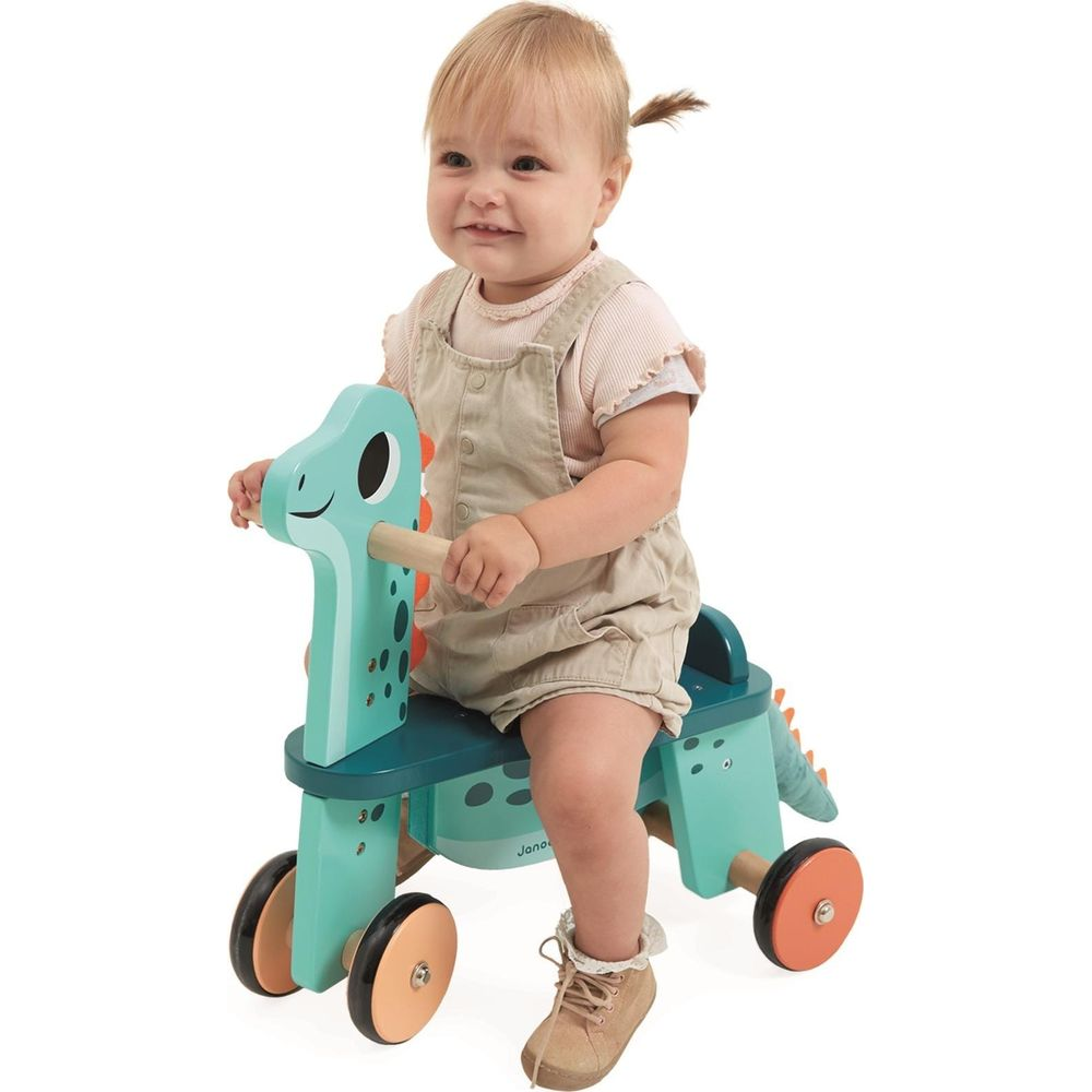 Ein Baby sitzt auf einem türkisblauen Dino-Laufrad mit Holzgriffen und Rädern.