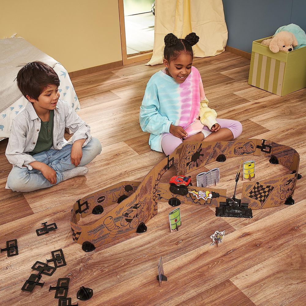 Zwei Kinder spielen mit einer Autorennbahn aus Pappe auf einem Holzboden.