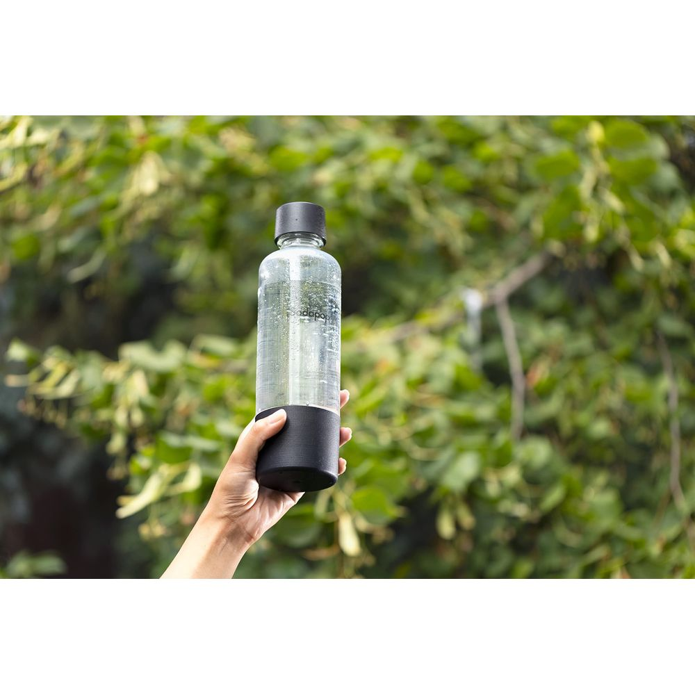 Eine Hand hält eine durchsichtige Flasche mit schwarzem Sockel vor grünem Laub.