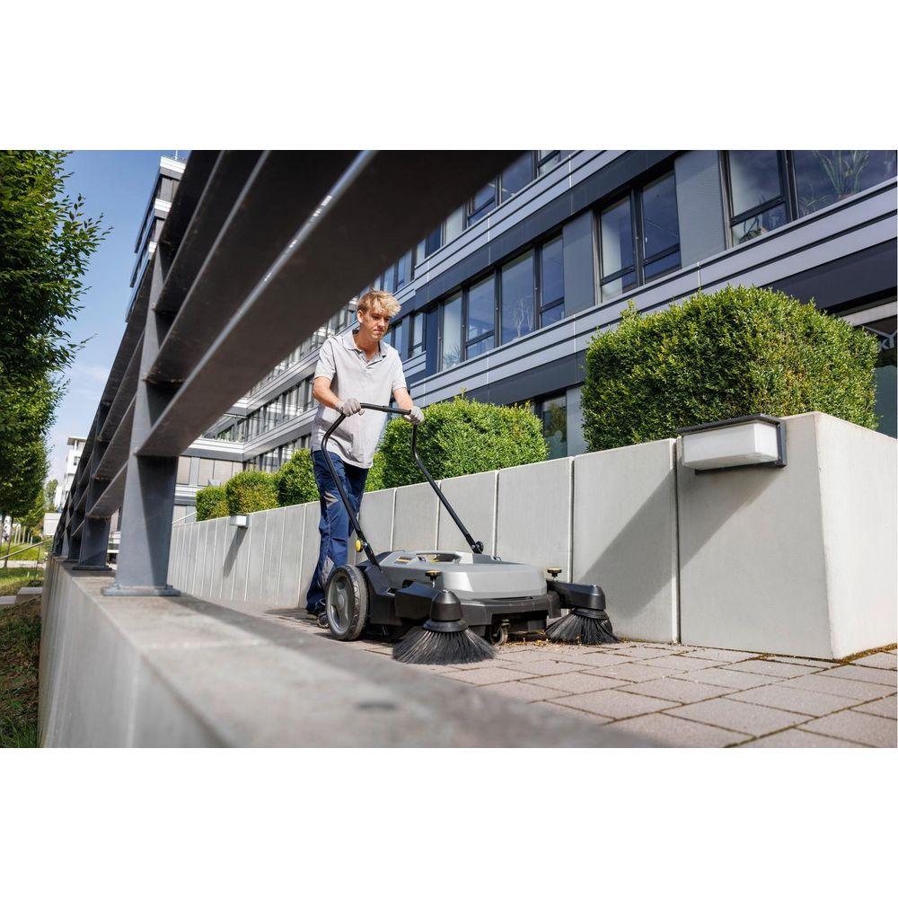 Mann schiebt Kehrmaschine auf gepflastertem Weg vor modernem Gebäude mit Büschen.