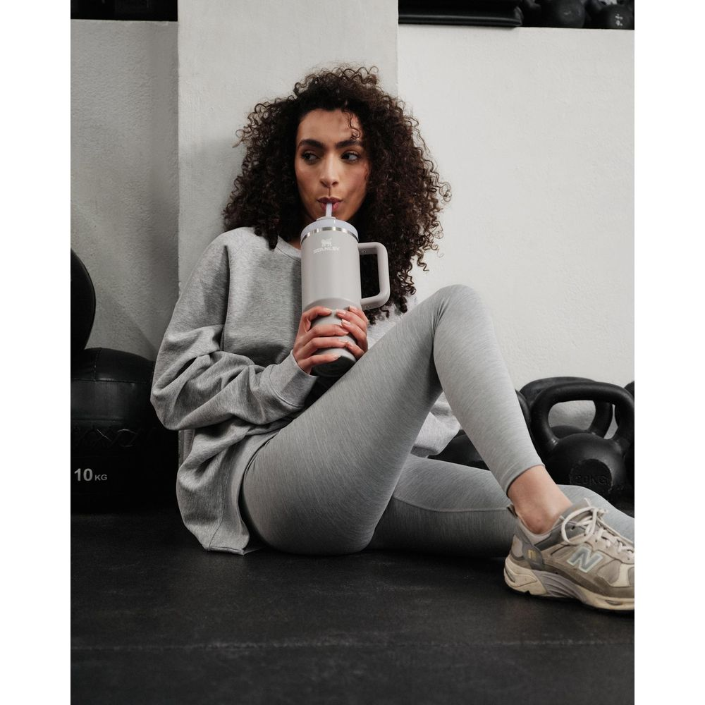Eine Frau mit lockigen Haaren trinkt aus einem grauen Becher in einem Fitnessstudio.