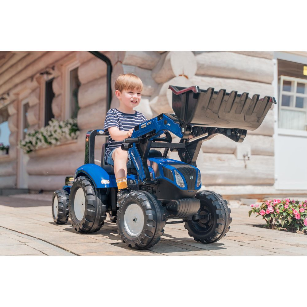 Ein kleiner Junge fährt einen blauen Spielzeugtraktor mit Lader vor einem Holzhaus.
